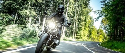 Mann mit schwarzem Helm fährt tagsüber Motorrad auf einer Landstraße durch den Wald