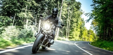 Mann mit schwarzem Helm fährt tagsüber Motorrad auf einer Landstraße durch den Wald